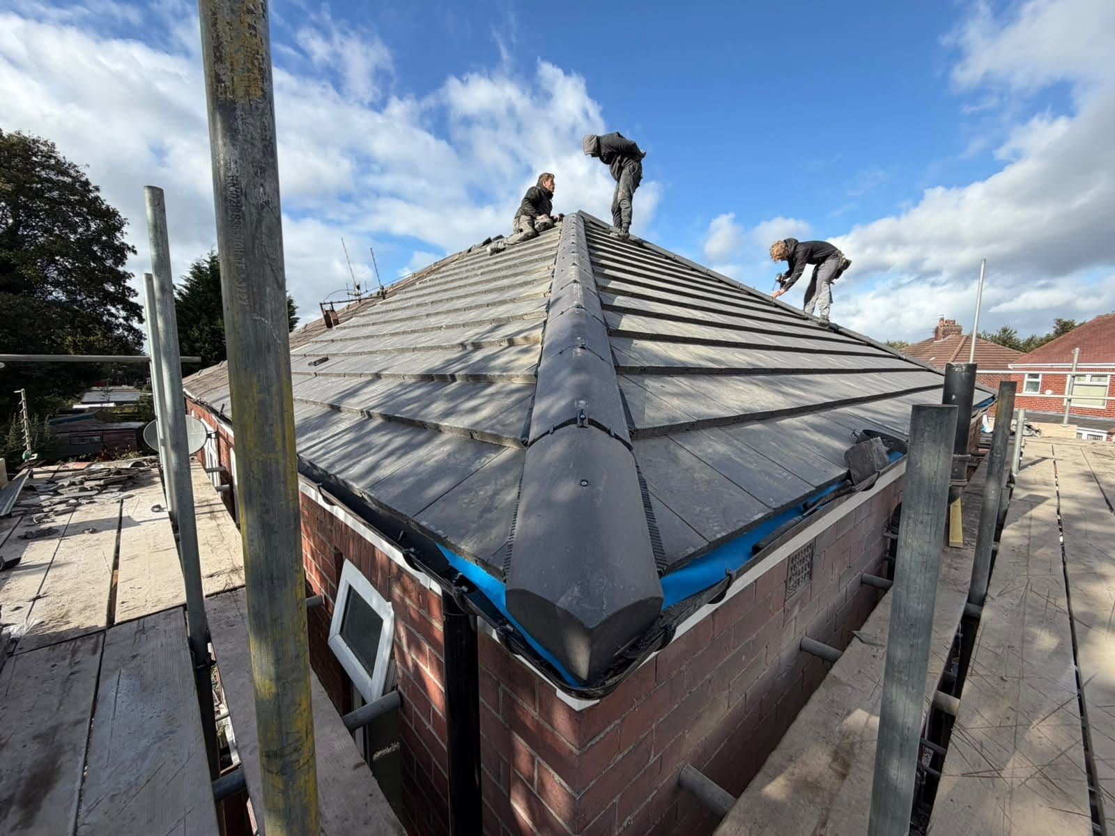 Three roofers working along the ridge and hips of a new grey tiled bungalow roof, with scaffold boards round the edge